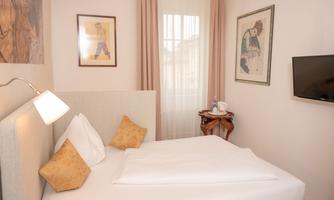 A cozy bedroom with a bright bed and decorative pillows. Artwork hangs on the wall, and a small table with a kettle is present.