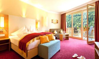 A bright hotel room with a large double bed, a cozy seating area, and a balcony overlooking nature. The carpet is red, and the room feels inviting and comfortable.