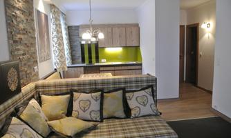 A modern living room with a cozy couch and stylish pillows. In the background, there is a functional kitchen with wooden cabinets and green accents.