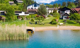 A peaceful landscape by the water with several houses and green gardens. In the background, mountains are visible under a clear sky.