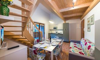 A cozy living space with a wooden staircase, a dining table, and colorful armchairs. The modern kitchen is well-equipped and lets in plenty of daylight.