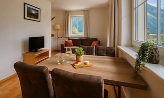 A cozy living room with a table, chairs, and a television. Large windows provide a view of the mountains in the background.