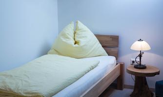A simple bedroom with a single bed and a yellow bedspread. Next to the bed is a nightstand with a lamp.