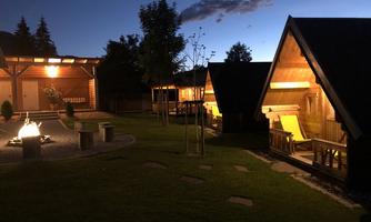 Cozy wooden cabins in a garden at night. A campfire and soft lighting create an inviting atmosphere.