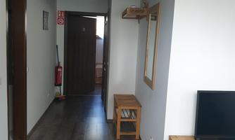 A bright corridor with wooden flooring and white walls. A mirror and a small shelf are hanging on the wall, while a television is placed on a wooden piece of furniture.