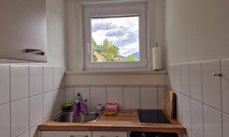 A simple kitchen with a sink and a small cooking area. Through the window, a view of the sky and the garden is visible.