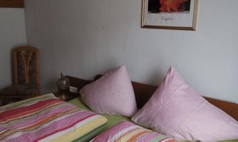 A cozy bedroom with a double bed and colorful bedspreads. A picture hangs on the wall and there is a wooden chair.