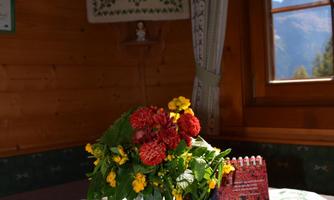 A cozy wooden table with a colorful bouquet in a white vase. In the background, there is a window overlooking the mountains and a statue.