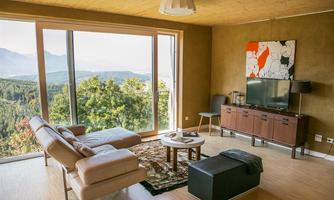 A modern living room with large windows and a view of the mountains. It is bright and elegantly furnished, with a comfortable sofa and a TV unit.
