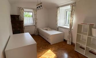 A bright room with parquet flooring and white furniture. There is a bed, a wardrobe, and a shelf, as well as windows with curtains.