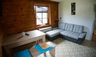 A cozy living room with a gray sofa and a wooden table. The walls are made of wood, and there is a window with blinds.