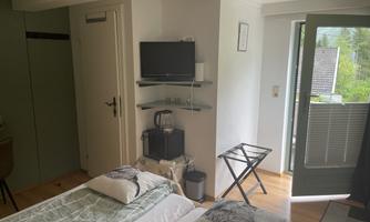 A simple room with two beds, a television, and a small seating area. The room has access to a balcony with a green view.