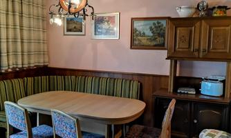 A cozy dining area with a round table and upholstered chairs. The walls are decorated with pictures and a lamp hangs from the ceiling.