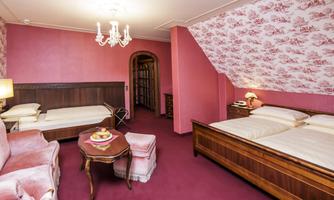 A cozy bedroom with two beds and a seating area. The walls are decorated in a pink pattern, and the furnishings are classic and inviting.