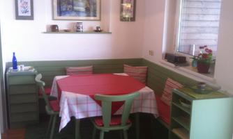 A cozy dining area with a red table and green chairs. The walls are light and there are windows looking outside.