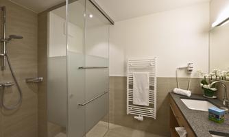 A modern bathroom with a glass shower and elegant tiles. The washing area is well equipped, with plants and a towel holder.