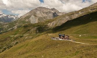 A charming mountain cabin in a vast, green landscape. In the background, majestic mountains rise under a partly cloudy sky.