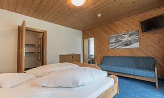 A cozy room with wooden ceilings, a double bed, and a blue sofa. A television is mounted on the wall, and there is a wardrobe and a small table.