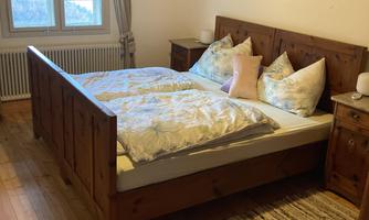 A cozy bedroom with a large wooden bed design and a soft carpet. The room has large windows and bright walls that create an inviting atmosphere.