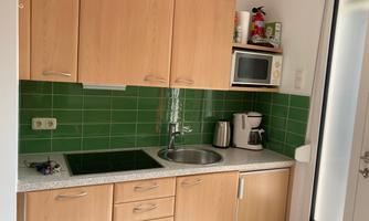 A modern kitchen with wooden cabinets and green tiles. It is equipped with a cooking stove, a sink, and kitchen appliances such as a microwave and a coffee maker.