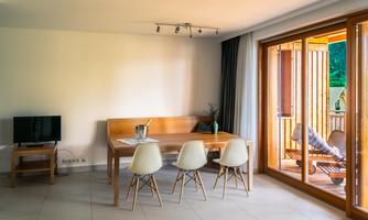 A modern living and dining room with a wooden dining table and white chairs. Large windows offer a view of a balcony with loungers.