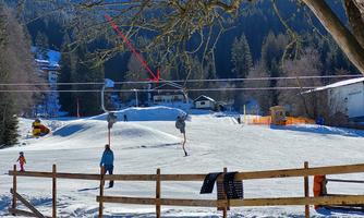 A ski resort with snow-covered ground and a ski lift. In the background, trees and buildings are visible.