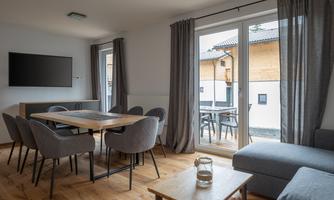 A modern living room with a dining table and comfortable chairs. Large windows let in a lot of light and offer a view of the terrace.