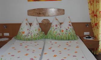 A cozy bedroom with a double bed and colorful cushions. A colorful painting hangs on the wall, and the curtains are also vibrantly designed.