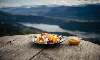 A plate of colorful food is on a wooden table. In the background, there is a beautiful mountain landscape with a body of water.