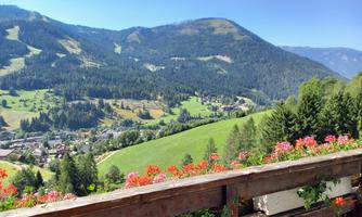 A calm mountain landscape with green meadows and lush trees. In the foreground, colorful flowers can be seen on a balcony.