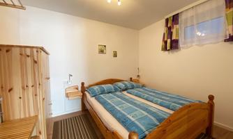 A simple bedroom with a wooden bed and colorful pillows. The walls are light, and there are a few pictures and a wooden wardrobe.