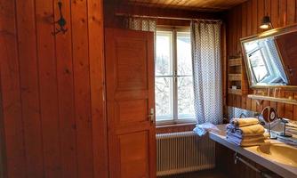 A cozy bathroom with wooden walls and a large window. Fresh towels are laid out on the washbasin and a mirror reflects the light.