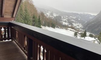 A balcony with a wooden railing overlooking a snowy mountain landscape. In the distance, forested hills and houses are visible.