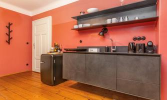 A modern kitchen with a gray kitchen unit and shelves on the wall. The walls are painted in a warm red tone, and the floor is made of wood.