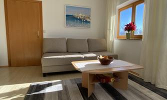 A bright living room with a gray sofa and a wooden table.  
On the table, there is a bowl of fruit, and there is a window with a view.