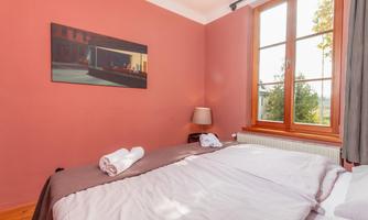 A cozy bedroom with pink walls and a large window. Fresh towels are laid on the bed, and a picture is hung on the wall.