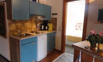 A small kitchen with blue cabinets and a stove. On the right, there is a glimpse into a bright room with a window and furniture.