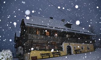 A charming wooden house in the snow, surrounded by falling snowflakes. The windows are lit, creating a cozy atmosphere in the wintry night.