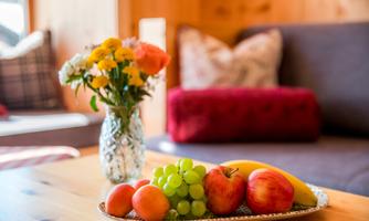 A table with a bowl full of fresh fruits and a vase with colorful flowers. In the background, a cozy living room with a sofa can be seen.