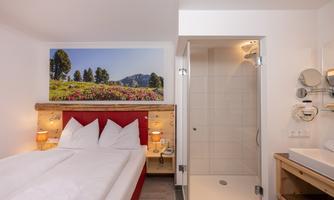 A modern hotel room with a double bed and a beautiful picture of a landscape on the wall. On the right, a bathroom with a shower is visible.