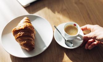 A fresh croissant on a plate next to a small cup of coffee. The sun is shining weakly on the table.