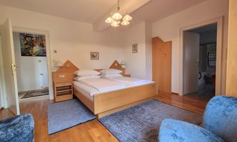 A cozy bedroom with a double bed and wooden furniture. There are two blue armchairs and a warm wooden floor.