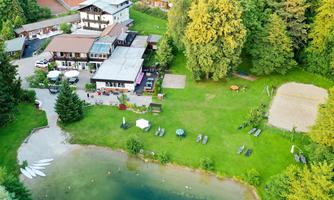A view of the trout farm amidst trees, surrounded by a meadow and a small swimming lake. There are terrace furniture and a sand area for recreational activities.