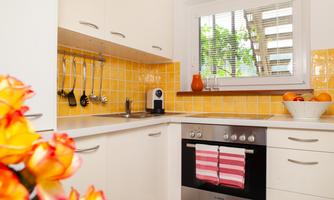 A modern kitchen with a bright, friendly design and yellow tiles. In the foreground, fresh flowers and a bowl of fruit are visible.