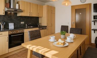 A modern kitchen with wooden cabinets and a dining table. On the table are cups and a small plant.