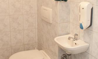 A simple bathroom with white tiles and a toilet. There is a small sink, paper towels, and a waste bin.