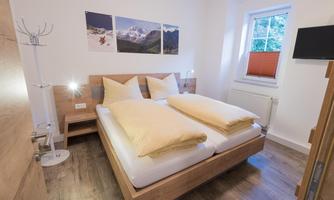 A cozy bedroom with a double bed and large, soft pillows. Pictures of mountains hang on the wall, and the room has plenty of natural light.
