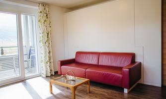 A cozy living room with a red sofa and a small table. Large windows provide plenty of daylight and a view outside.