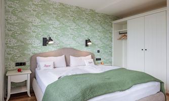 A modern bedroom with a large bed and fresh green accents. The walls are decorated with a stylish pattern and there is a white wardrobe.