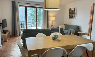 A modern living room with a dining area and a large window. There is a cozy sofa, a television, and a warm atmosphere.